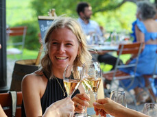 Three women toast with sparkling white wine, smiling and seated outdoors in a friendly, relaxed atmosphere. Gourmet appetizers are on the table, while others enjoy the evening in the background. The scene conveys joy, togetherness, and good taste.