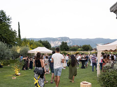 In a large garden surrounded by olive trees, many people gather for an outdoor event. White tents and tall tables create an elegant and relaxed atmosphere. In the distance, mountains can be seen, while soft lights illuminate the scene. The mood is convivial, blending nature, wine, and music.