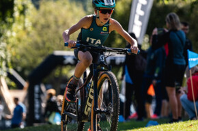 A child pedals his bike with determination on the beach of Lake Molveno. He wears a helmet, sunglasses and a green uniform, with ITA written on his chest.