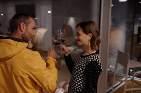 degustazione in cantina Rotari Un uomo e una donna  stanno alzando i loro calici di vino rosso, sorridendosi a vicenda. Si trovano all'interno di una cantina moderna con pareti di vetro e pavimenti in legno. Dietro l'uomo, sono visibili delle botti di vino accatastate in un'altra stanza. L'atmosfera è intima e l'illuminazione è soffusa.