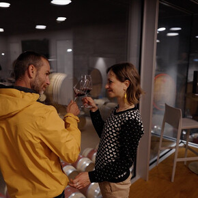 degustazione in cantina Rotari Un uomo e una donna  stanno alzando i loro calici di vino rosso, sorridendosi a vicenda. Si trovano all'interno di una cantina moderna con pareti di vetro e pavimenti in legno. Dietro l'uomo, sono visibili delle botti di vino accatastate in un'altra stanza. L'atmosfera è intima e l'illuminazione è soffusa.