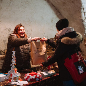 The photo shows the Christmas craft markets inside a vaulted passage in Faedo. In the center of the photo, there is a young woman who has bought something and is collecting her paper bag from the vendor.