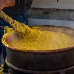 La foto cattura il primo piano di un paiolo colmo di polenta mentre un uomo mescola con un grande bastone in legno