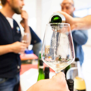 Calice di vino bianco durante una degustazione, con persone sullo sfondo che assaggiano e dialogano.