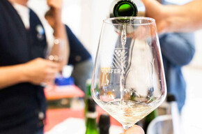 Calice di vino bianco durante una degustazione, con persone sullo sfondo che assaggiano e dialogano.