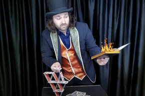 Nell'immagine si vede il mago mentre sfoglia un libro infuocato e costruisce un castello di carte