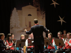 Un'orchestra è ritratta su un palco. Il direttore, di spalle, è al centro e dirige i musicisti seduti, la maggior parte dei quali indossa gilet rossi e camicie bianche. Gli strumenti includono clarinetti, sassofoni e una tuba. Sullo sfondo nero è appeso un telo raffigurante una chiesa bianca con un campanile e alberi innevati, affiancato da grandi decorazioni a forma di stella. L'atmosfera è quella di un concerto o di un'esibizione natalizia al chiuso.