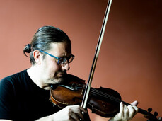 Un musicista suona il violino durante l'evento Sounds of Sauna, con un percussionista sullo sfondo in un'atmosfera soffusa.