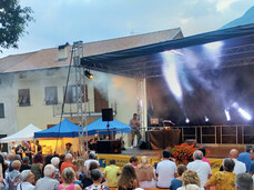 The image shows an outdoor community event with a stage where a person is operating equipment, possibly a DJ or presenter. A crowd of people, mostly middle-aged and older, watches the performance while seated or standing in an open area with a reddish floor. The setting appears to be in a small town or village with mountains visible in the background, and there are some market tents and colorful decorations visible, suggesting it might be a local festival or cultural celebration.