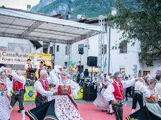  Un gruppo di persone, uomini e donne, esegue una danza tradizionale su un palco all'aperto con un pavimento rosso. I ballerini indossano abiti folcloristici dai colori vivaci: gilet rossi, camicie bianche e gonne ampie con motivi floreali che si alzano durante il movimento. Dietro di loro si trova il palco principale coperto, con uno striscione di una cassa rurale e attrezzatura audio. La scena si svolge in un cortile circondato da edifici chiari e alberi, suggerendo un festival o una celebrazione di paese.