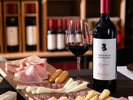 A close-up of a rich platter of cold cuts, cheeses, and breadsticks, accompanied by a bottle of Teroldego Rotaliana Riserva from Cantina Rotaliana and a full glass showcasing the intense red color of the Teroldego. The scene is set inside the winery’s Wine Shop, with shelves of bottles visible in the background.
