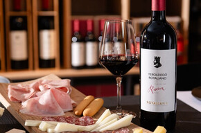 A close-up of a rich platter of cold cuts, cheeses, and breadsticks, accompanied by a bottle of Teroldego Rotaliana Riserva from Cantina Rotaliana and a full glass showcasing the intense red color of the Teroldego. The scene is set inside the winery’s Wine Shop, with shelves of bottles visible in the background.