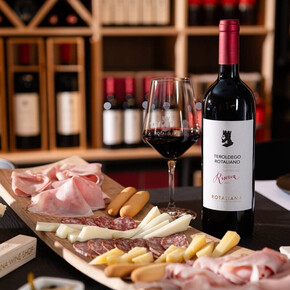 A close-up of a rich platter of cold cuts, cheeses, and breadsticks, accompanied by a bottle of Teroldego Rotaliana Riserva from Cantina Rotaliana and a full glass showcasing the intense red color of the Teroldego. The scene is set inside the winery’s Wine Shop, with shelves of bottles visible in the background.