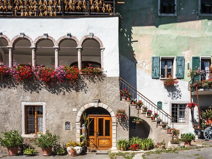 A view of San Lorenzo in Banale on a sunny day. The red and purple spots of the many pots of flowers, perhaps geraniums, stand out in the picture: they are hanging from the windows and the edge of a terrace surrounded by arches, leaning on the steps and on a small balcony. The sunshine and these bright colours convey a sense of cheerfulness.